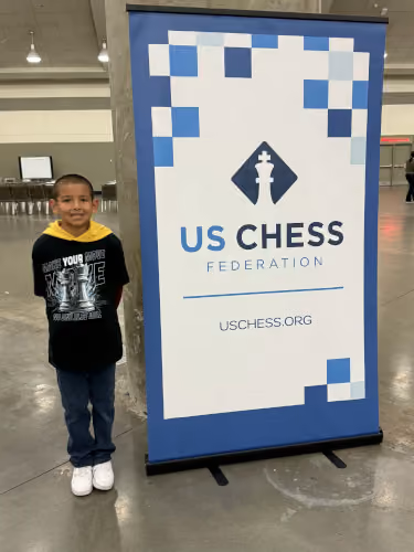 Young RGV Chess player competing at the US Chess National Championship, posing next to an official US Chess Federation event banner.