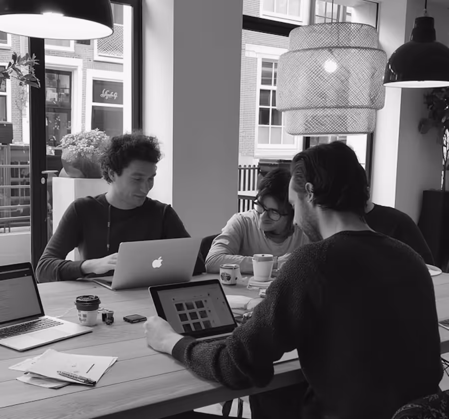 Four people sitting around a wooden table working on laptops and a tablet in a room with large windows and modern hanging lamps.