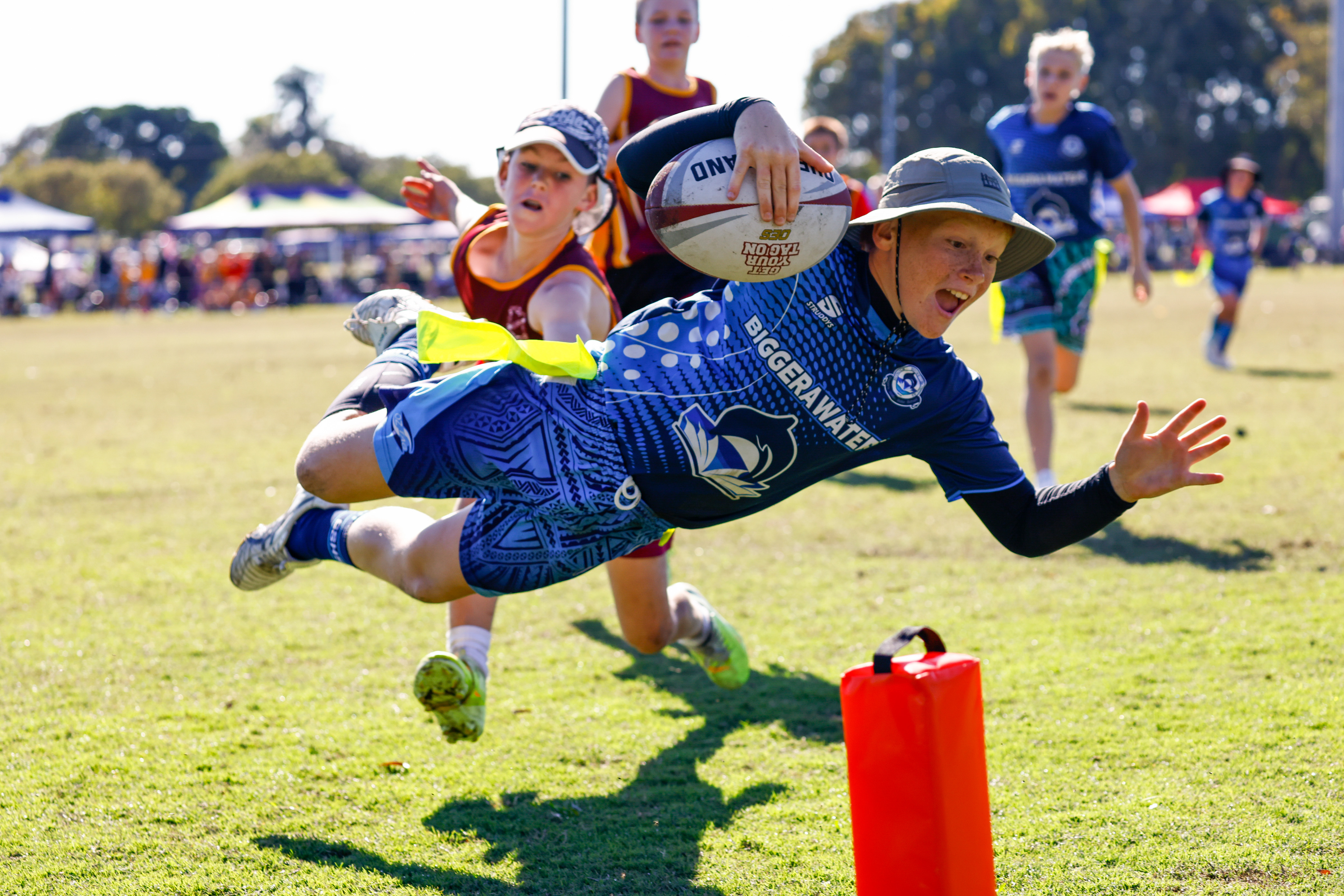 Junior tag rugby player in a blue jersey diving to score a try with the ball held out while a defender reaches for his belt flag