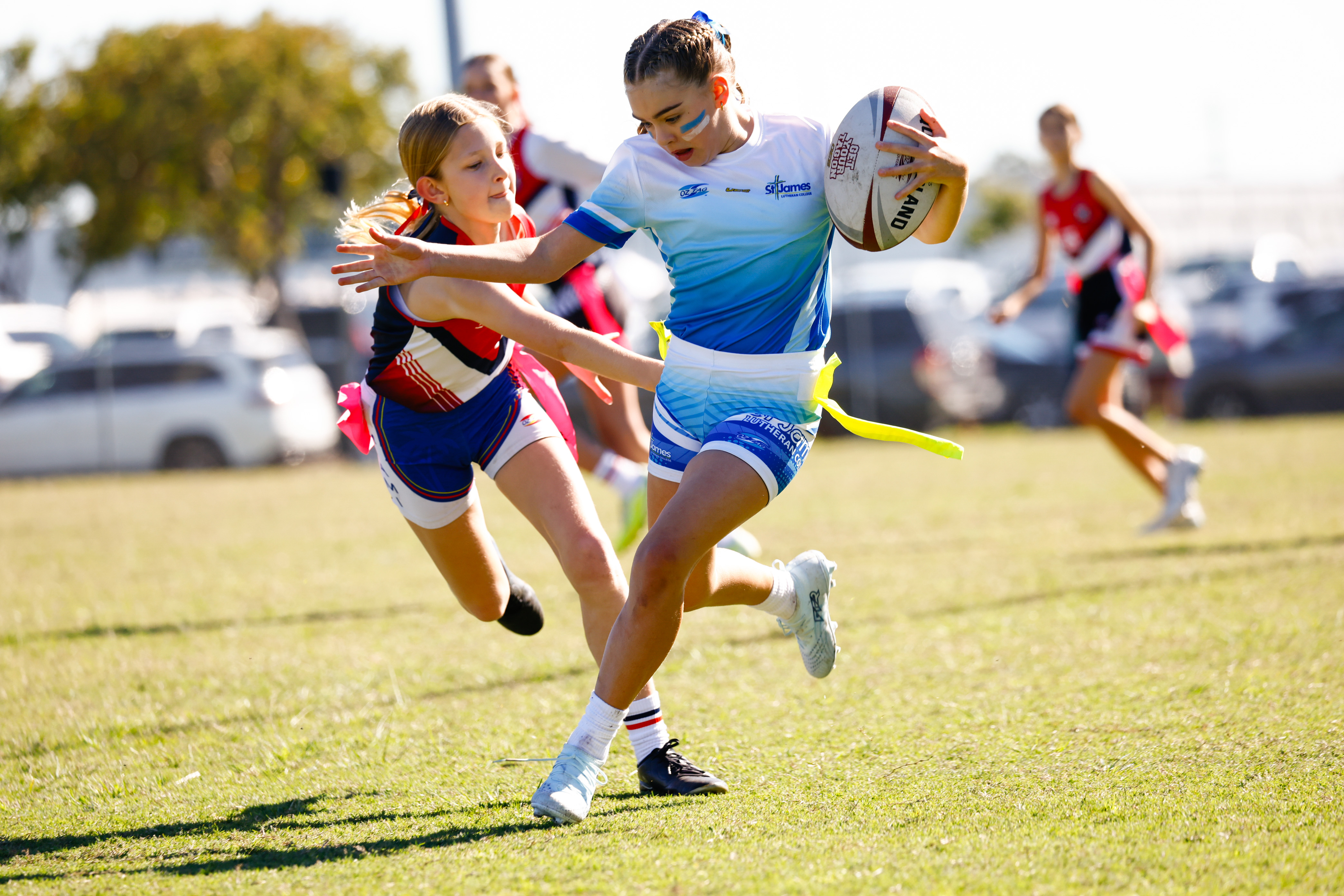 Girl in a light blue jersey running with the ball while a defender reaches for her flag in a junior touch football game