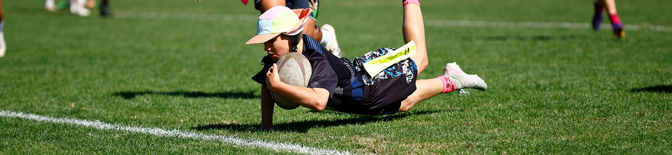 Junior tag rugby player in a blue jersey diving to score a try with the ball held out while a defender reaches for his belt flag