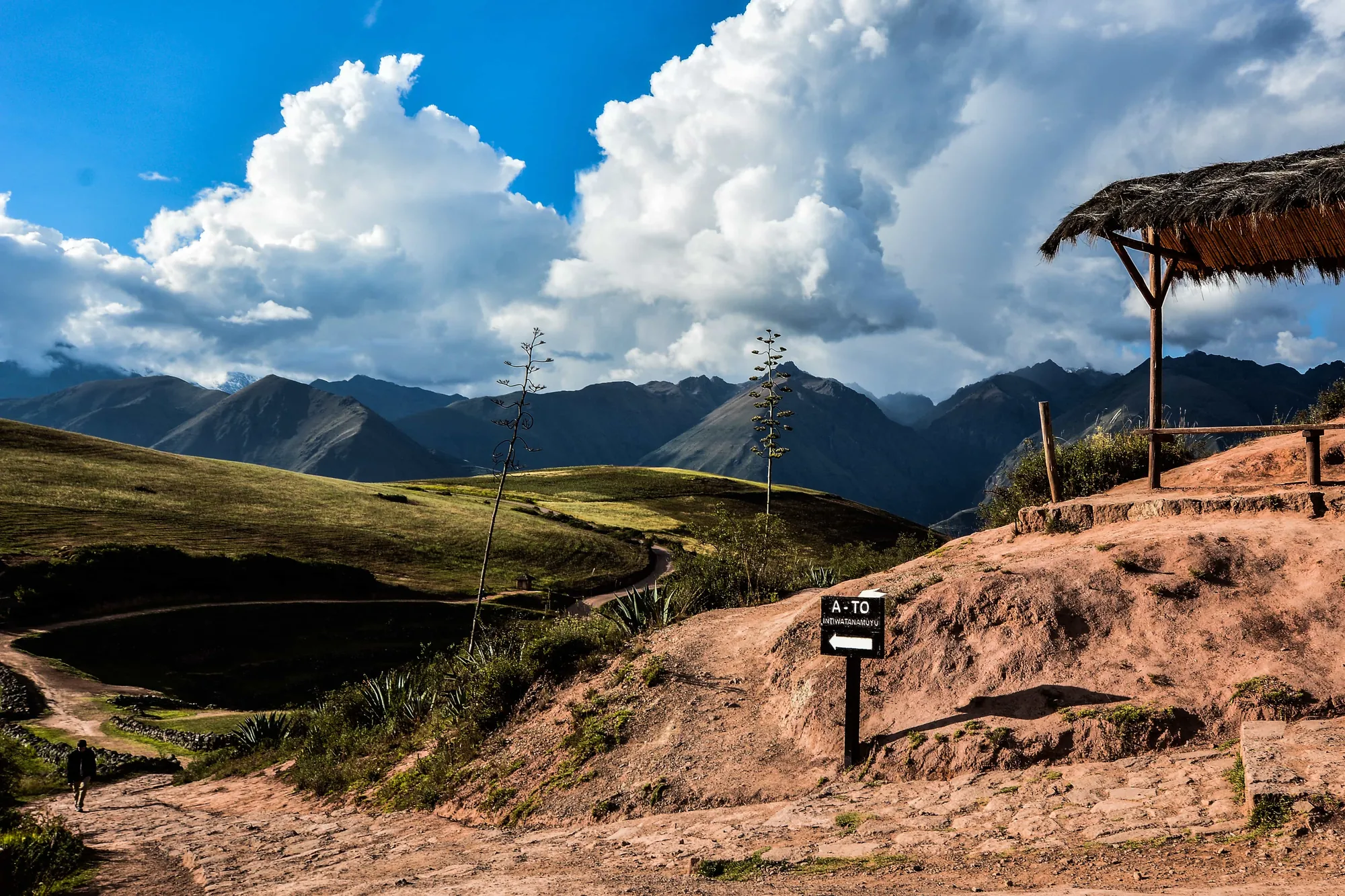 Green Andean valley with a thatched shelter and a trail sign under heavy cloud