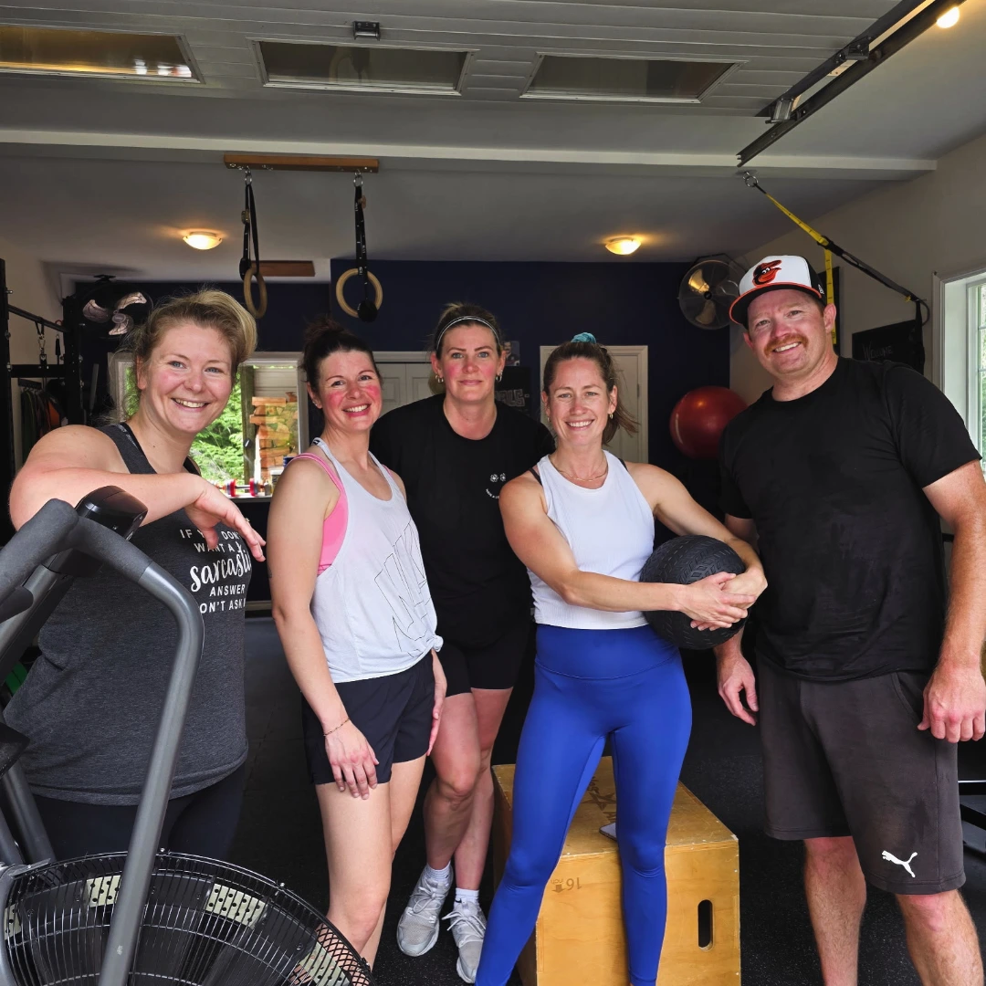Five adults, four women and one man, smiling in a gym setting; one woman in blue leggings holds a black medicine ball while standing on a wooden plyometric box with gym equipment and rings hanging in the background.
