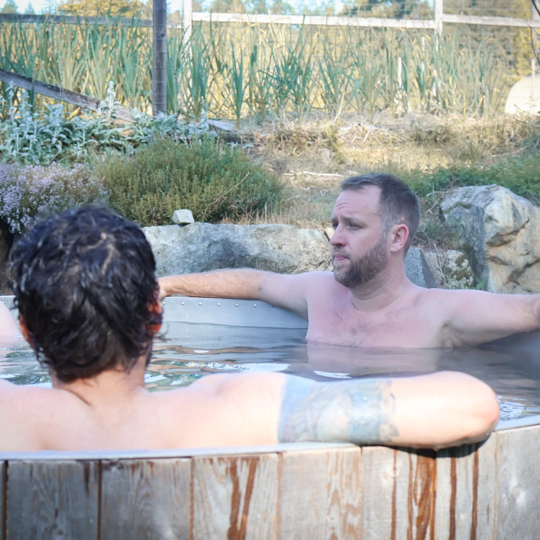 Two men relaxing in an outdoor wooden hot tub surrounded by greenery and rocks, with one man facing the camera and the other showing his back and tattooed arm.