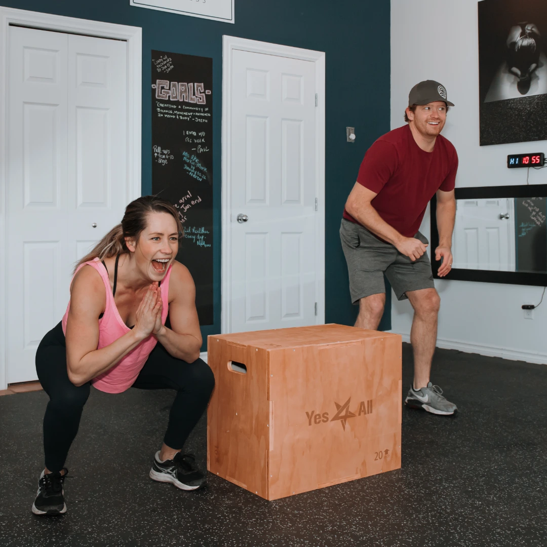 A woman in a pink tank top and black leggings squats with hands clasped in front of her in a gym setting, while a man in a red shirt, gray shorts, and a gray cap stands nearby smiling, with a wooden workout box labeled 'Yes4All' between them.