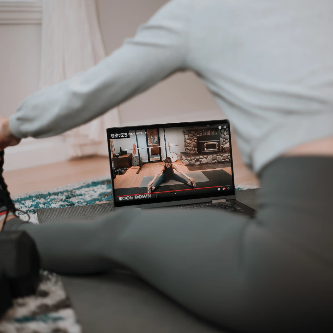 Person stretching on a yoga mat in front of an open laptop displaying a workout video with a woman stretching indoors near a stone fireplace.