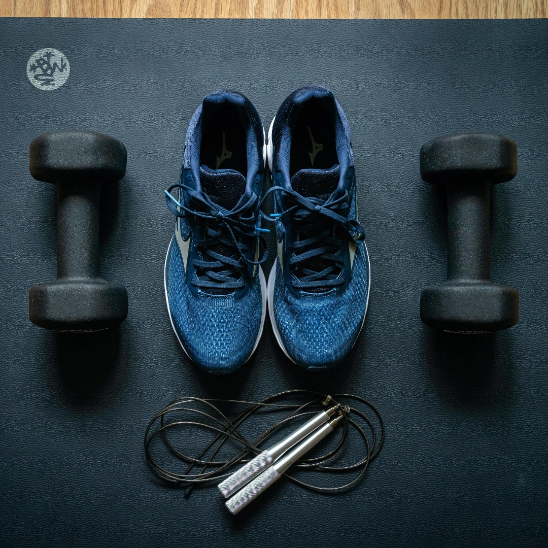 Pair of blue athletic shoes placed on a black exercise mat, flanked by two black dumbbells and a silver-handled jump rope below the shoes.