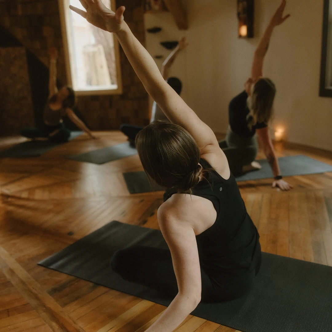 Four women seated on yoga mats indoors in a wooden-floored room, stretching one arm up and to the side in a yoga pose.