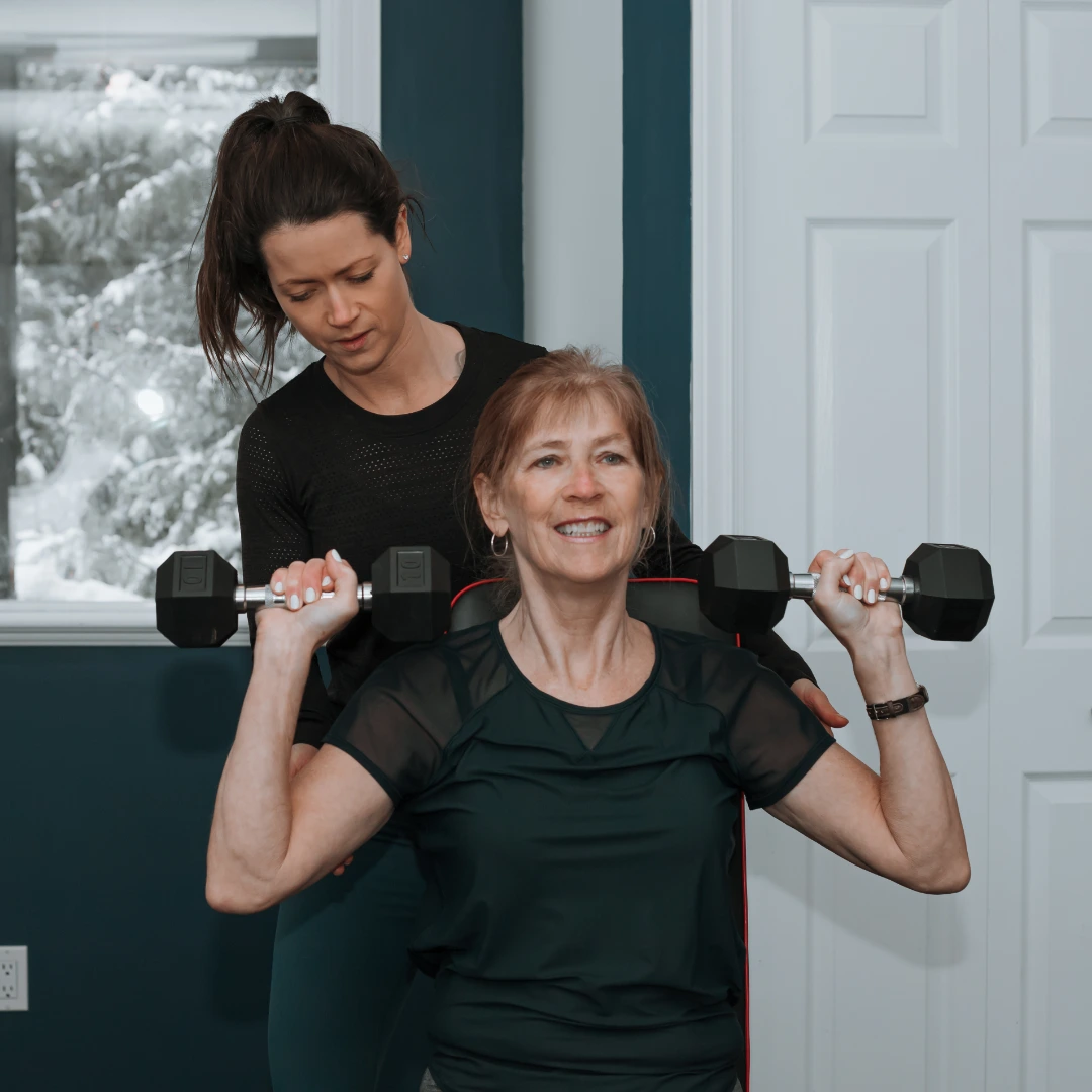 An older woman sits on a workout bench lifting dumbbells with both hands while a younger woman stands behind her, providing support and guidance during the exercise inside a room with a window showing snow outside.