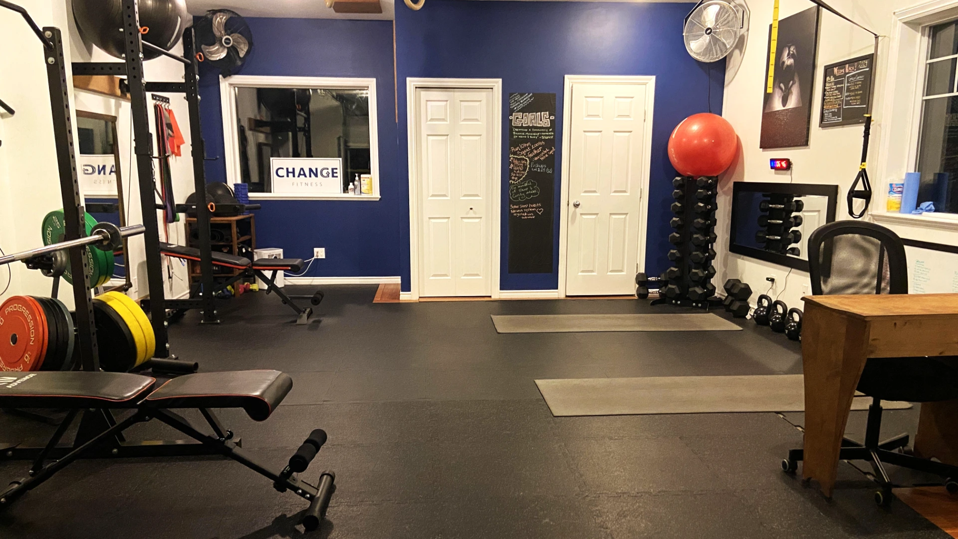 A fully equipped private fitness studio featuring a weight rack, exercise benches, dumbbell tower, kettlebells, and workout mats set up on black flooring.