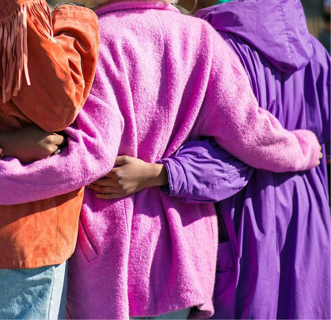 Three people in orange, pink, and purple jackets with arms around each other from behind.