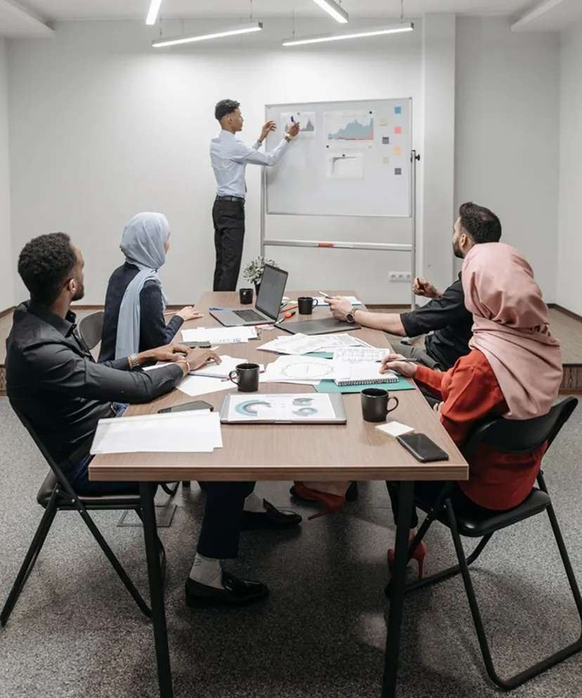 A diverse group of five people in a meeting room with a wooden table, looking at a man standing by a whiteboard presenting charts and graphs.