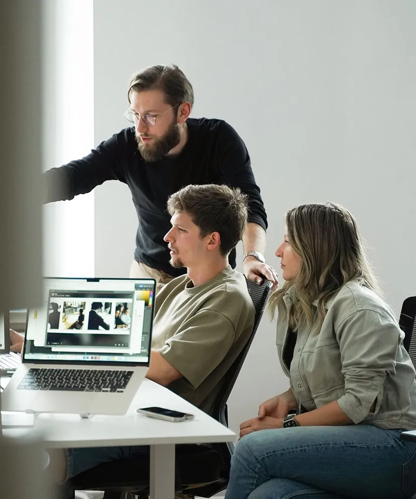 Three coworkers in a modern office collaborating around a laptop displaying a photo editing screen, with one standing and pointing while the other two sit and attentively watch.