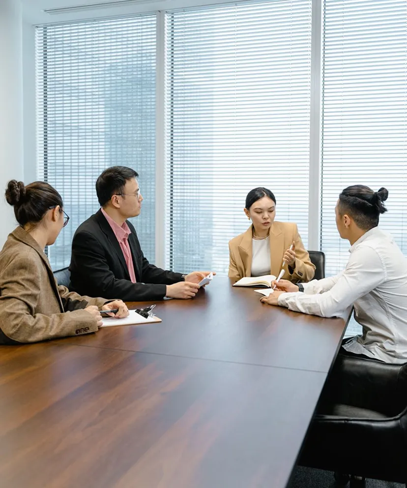 Four business professionals having a meeting in a modern office with large windows and blinds, seated around a wooden conference table reviewing documents and notes.