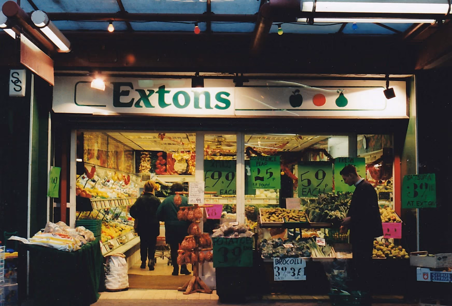 Exterior view of Extons fruit and vegetable shop with customers and price signs in the window.