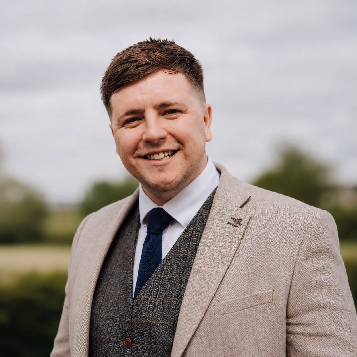 Smiling man in a beige blazer, gray vest, white shirt, and navy tie outdoors.