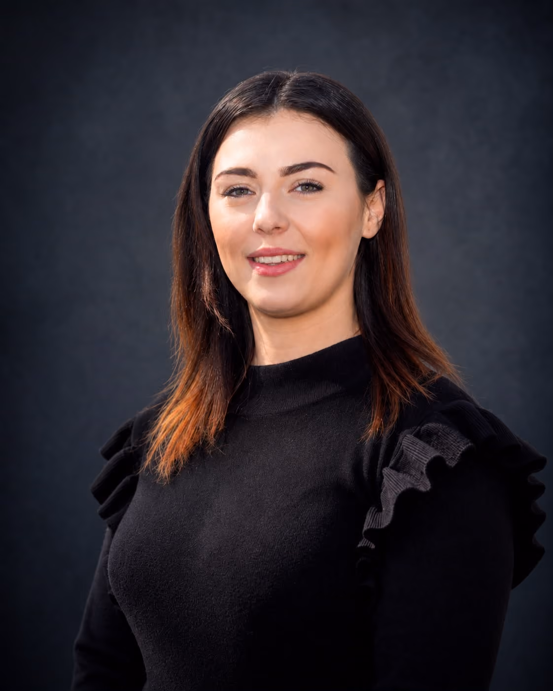 Smiling woman with dark hair in a black top with ruffled shoulders against a dark background.