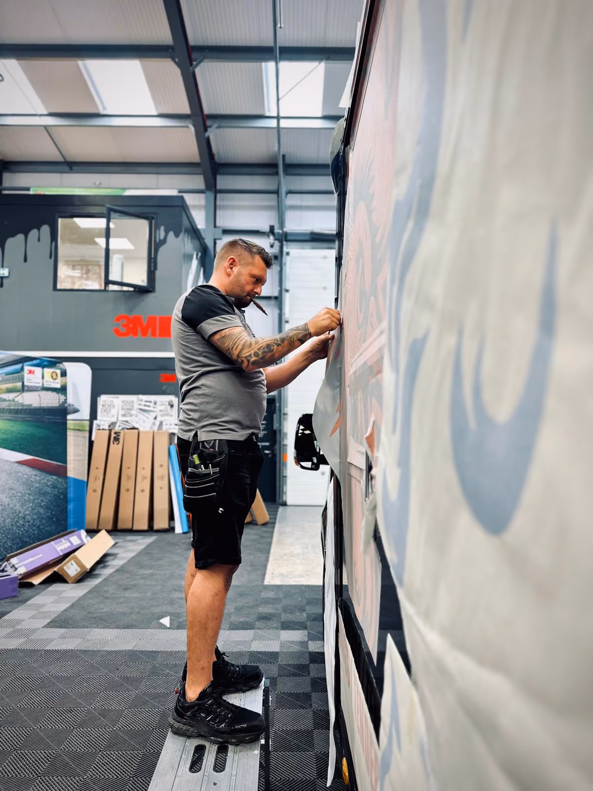 Man with tattooed arm standing on stool applying large decal to vehicle side in workshop.