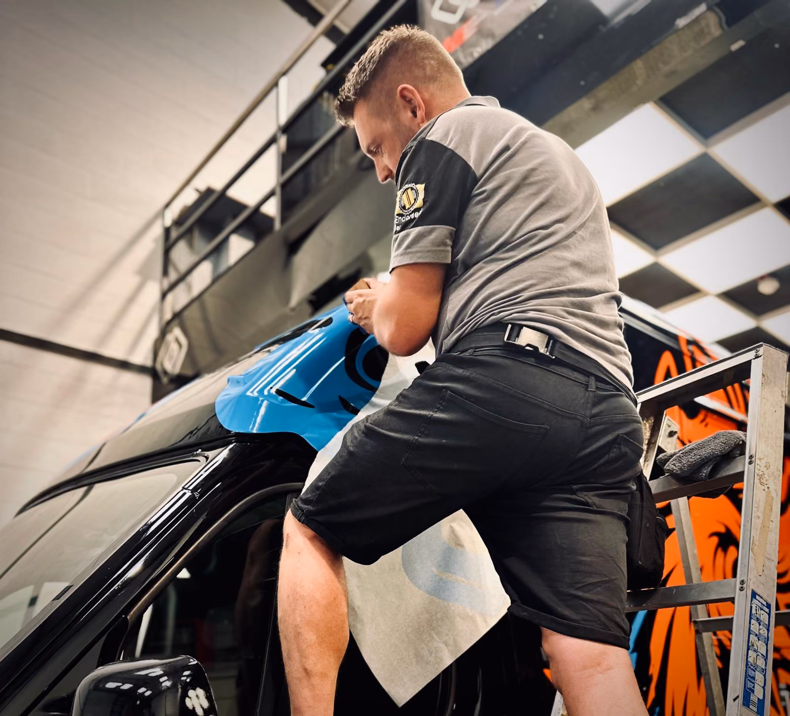 Man in gray shirt applying blue vinyl wrap to a black vehicle using a ladder indoors.