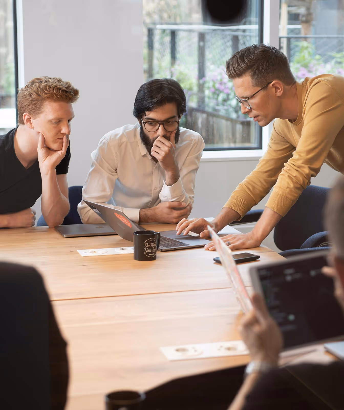 Three men in a meeting room focused on a laptop screen, with one standing and pointing at the laptop keyboard while two others sit and watch attentively.