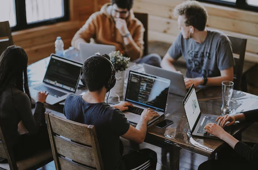 Group of young people working on laptops around a wooden table in a casual office setting.