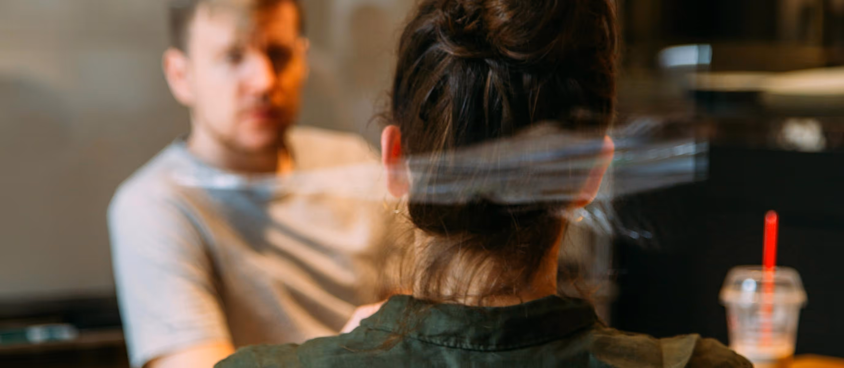 Two people having a conversation across a table in a softly lit room, with the woman’s back toward the camera and the man facing forward.