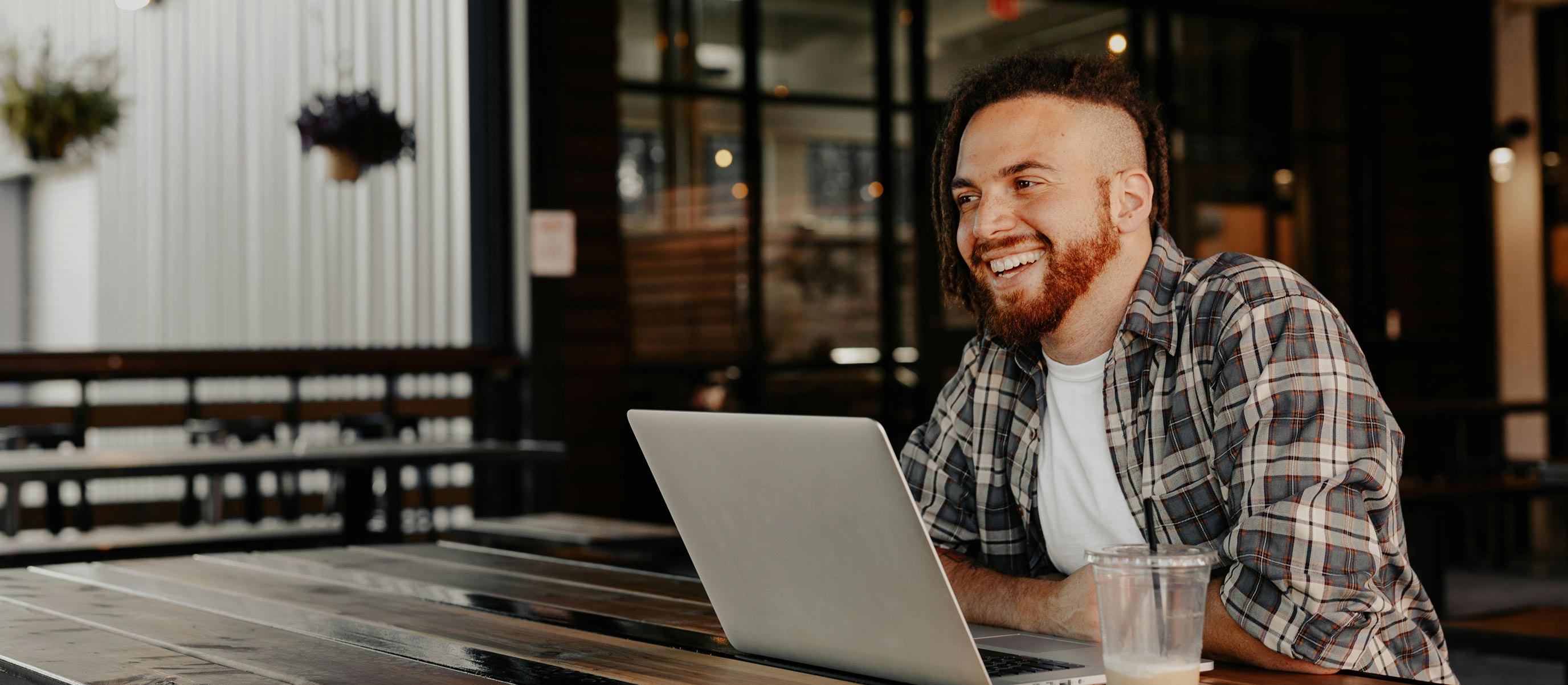 Smiling man with a beard and dreadlocks using a laptop at a wooden table in a café.