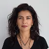 Woman with dark curly hair wearing a black top and layered necklaces, looking confidently at the camera against a white background.