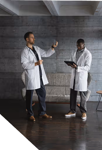 Two doctors in white coats with stethoscopes, one examining a syringe and the other checking a tablet, standing in a modern room with a gray couch.