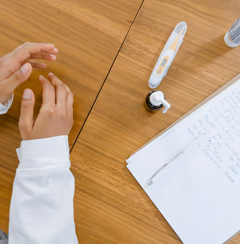 A person wearing a white lab coat with hands near a digital thermometer, medicine bottle, and handwritten notes on a wooden table.
