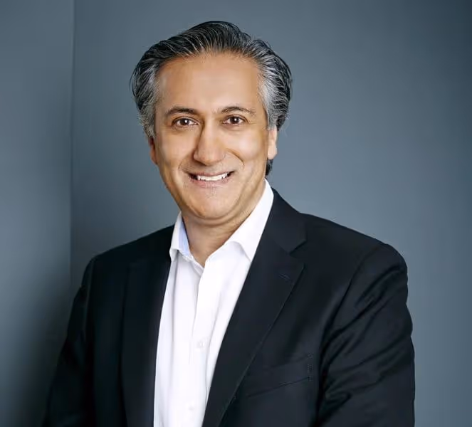 Man with gray hair wearing a black blazer and white shirt smiling against a gray background.