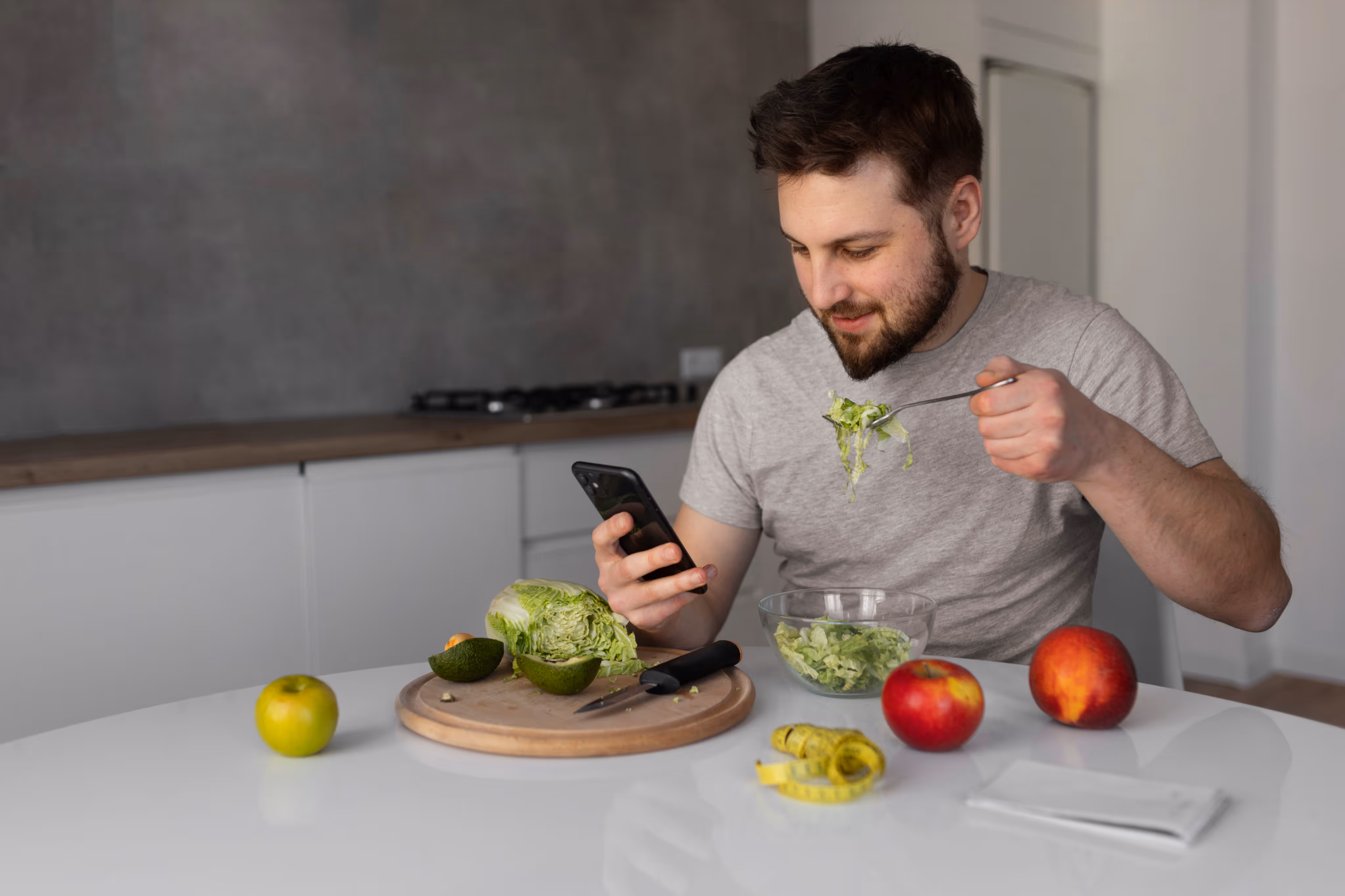 Man in a gray t-shirt eating salad while looking at his smartphone at a kitchen table with fruits and a measuring tape.