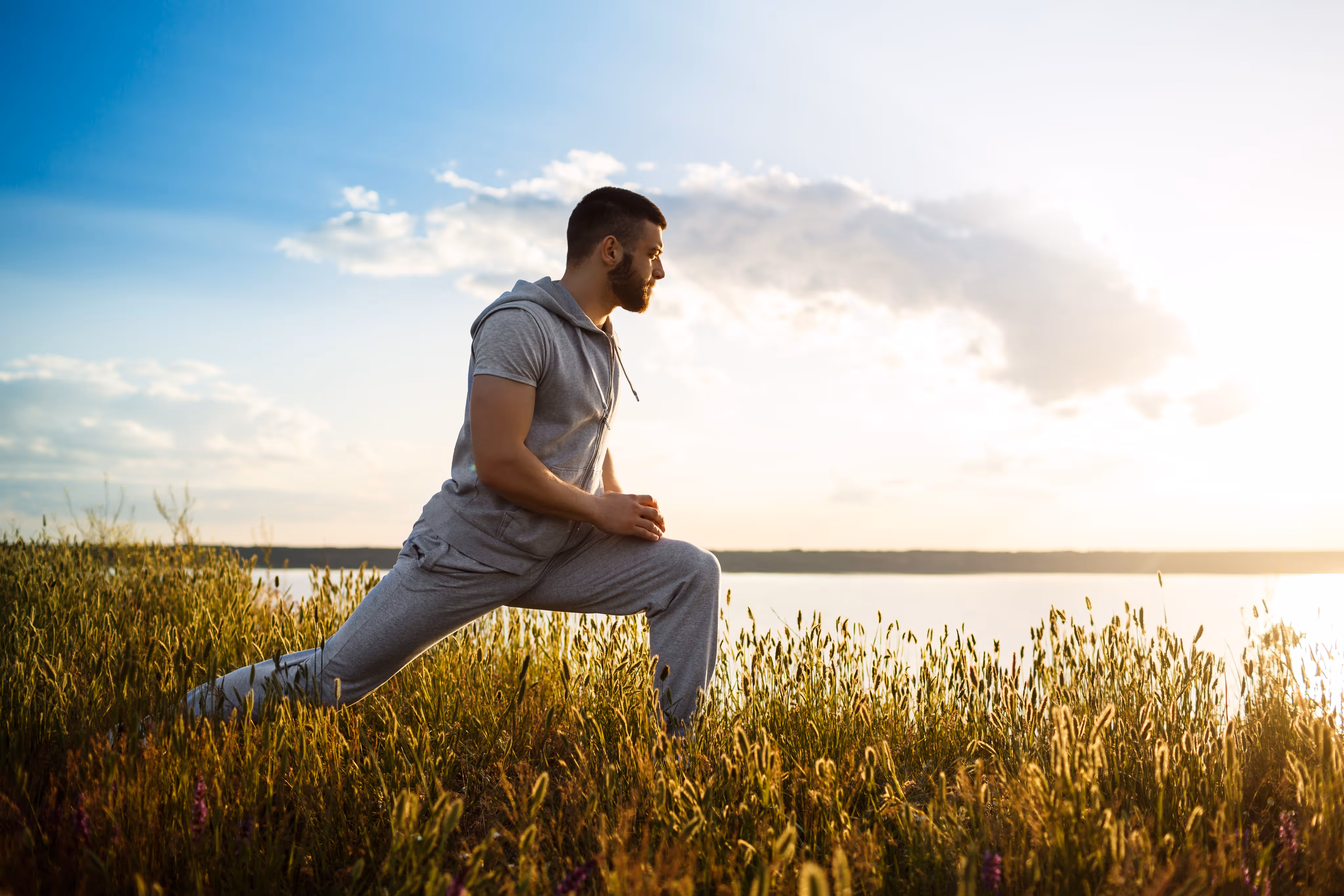 Man in gray athletic wear stretching his leg in a sunlit field near a body of water at sunset.
