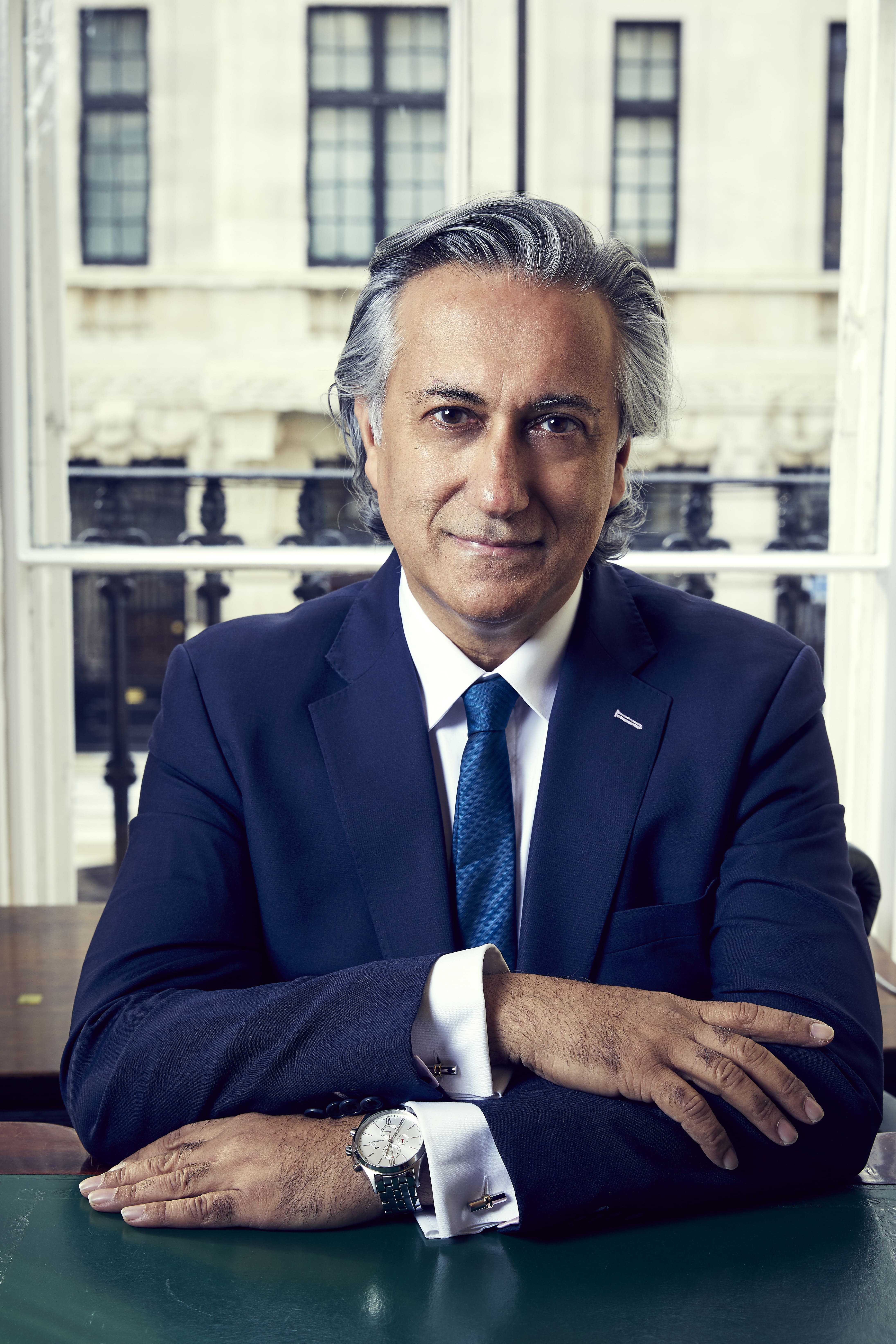 Middle-aged man with gray hair wearing a navy suit and blue tie seated at a desk with arms crossed, smiling slightly.