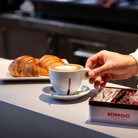 Kimbo kapućino sa latte art dekoracijom na šanku bara, pored svežih kroasana.