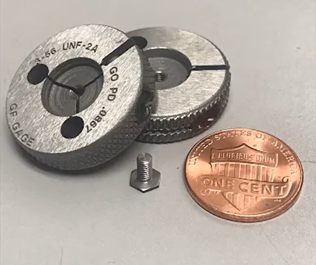 Two metal threaded gauges, a small bolt, and a U.S. penny for size comparison on a table.