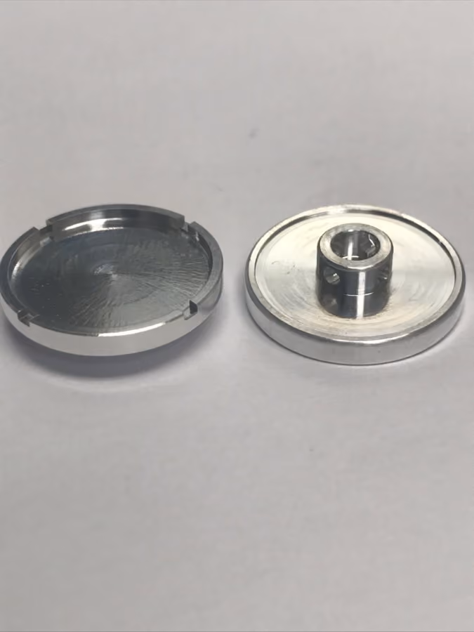 Two round metal parts on a white surface, one hollowed and the other with central raised hole and side holes.