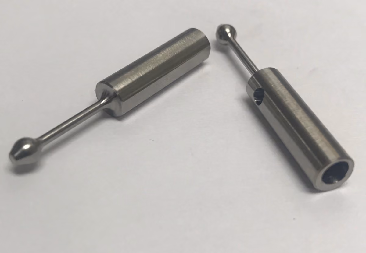 Two small cylindrical stainless steel metal parts with ball ends on rods lying on a white surface.
