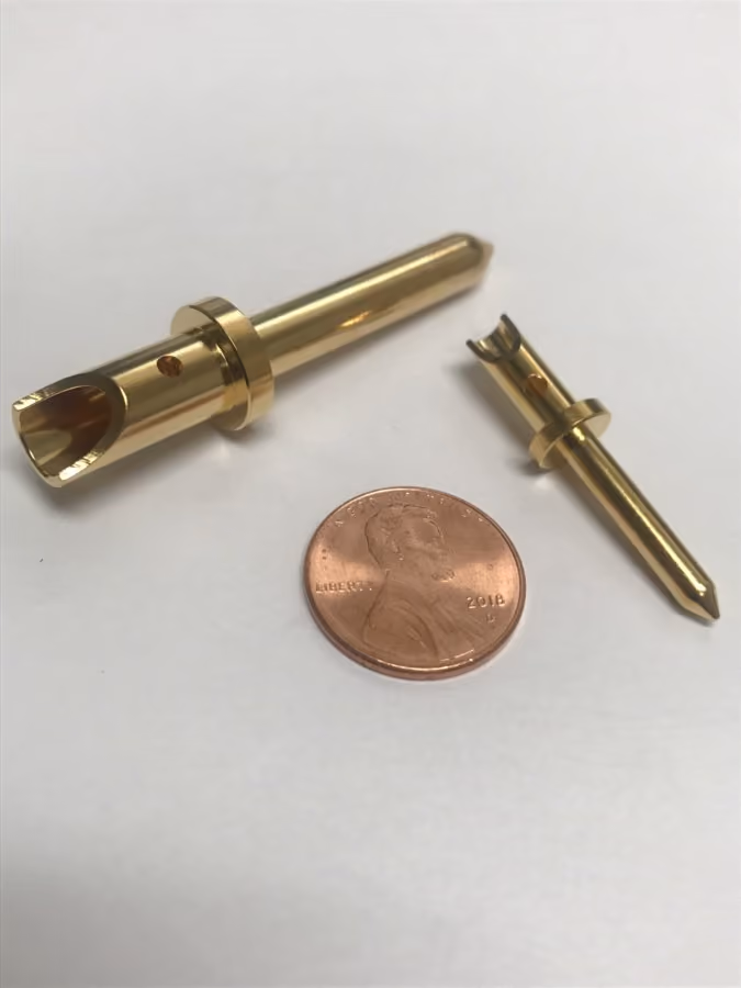 Two brass metal pins with a hole, next to a 2018 US penny for size comparison on white surface.