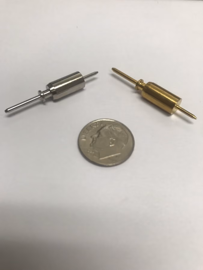 Two metal pins, one silver and one gold-colored, placed beside a U.S. dime on white surface.