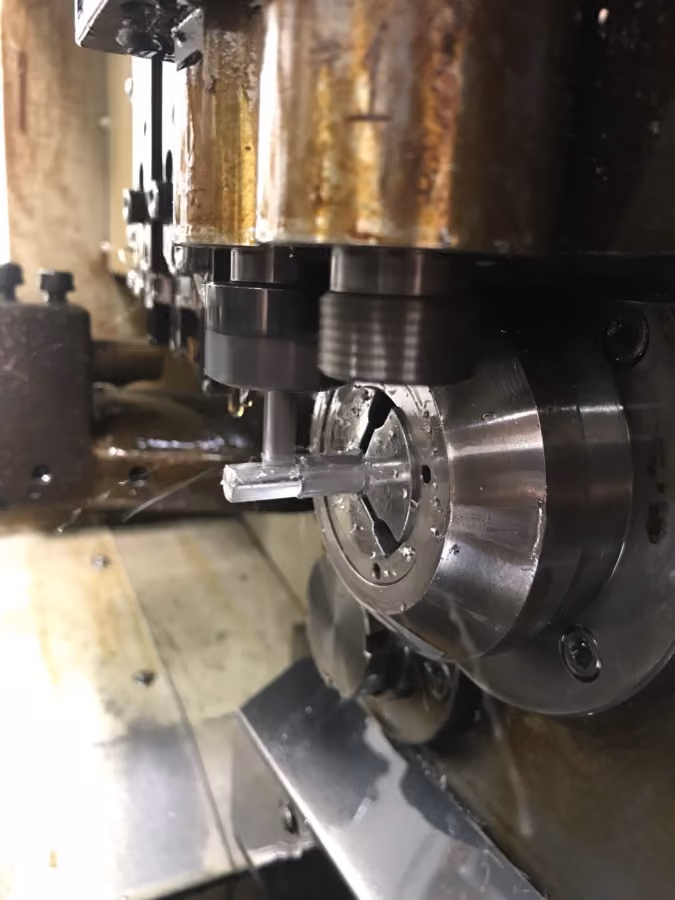 Close-up of a metalworking machine cutting a cylindrical metal piece with a vertical tool in an industrial setting.
