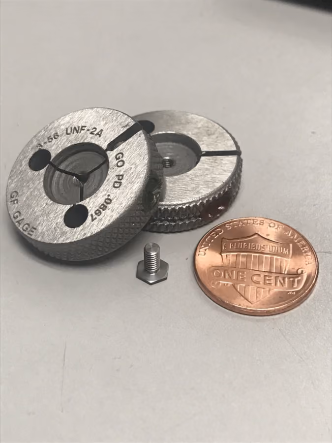 Two metal gauge tools, a small bolt, and a US one-cent coin on a gray surface.