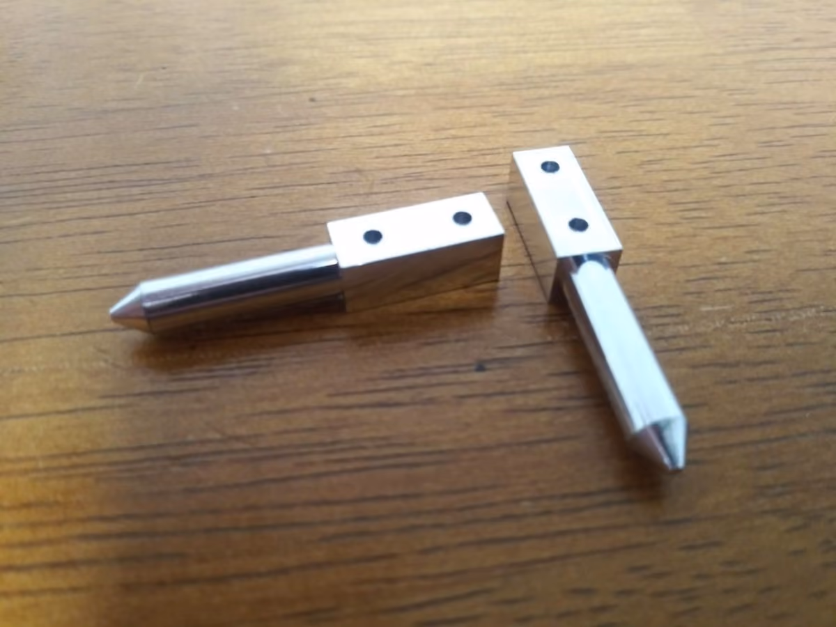 Two small metal parts with pointed ends and rectangular blocks with holes resting on a wooden surface.