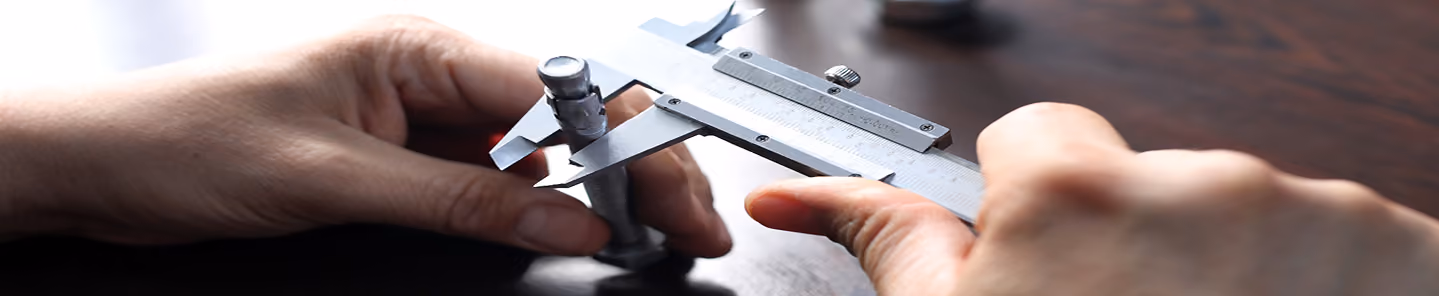 Hands using a caliper to measure the diameter of a metal bolt on a dark surface.