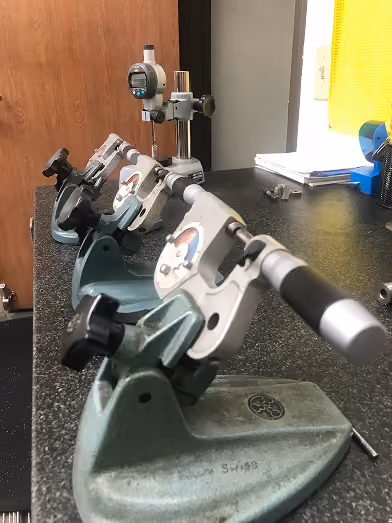 Micrometer tools with dials arranged in a row on a black speckled workbench near a wall.