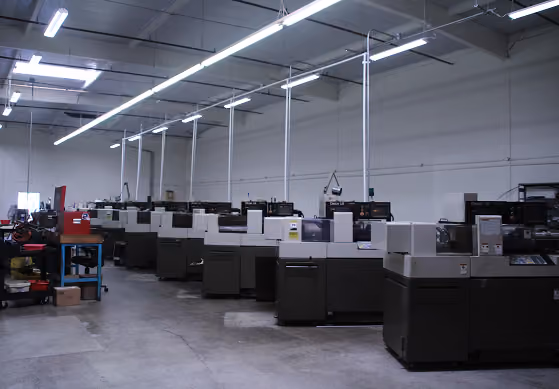 Inside a factory with a row of large industrial printing machines under long ceiling lights.