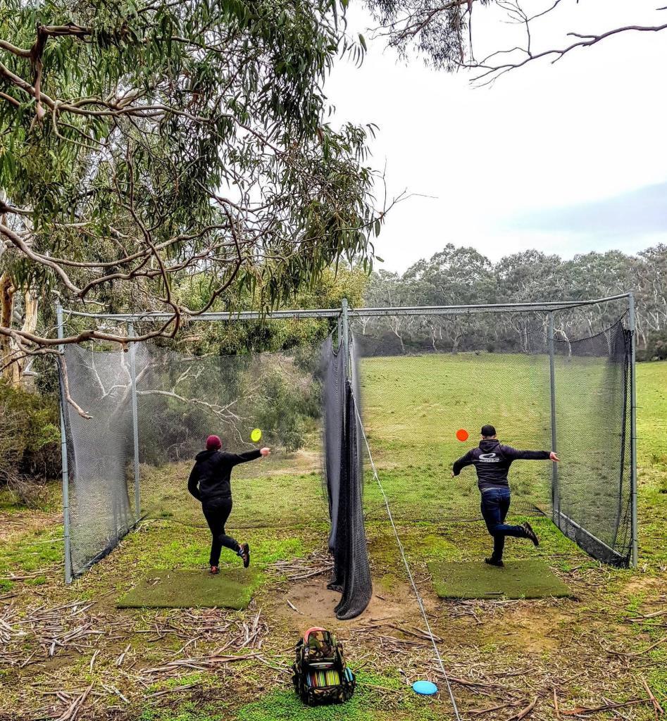 Two disc golfers teeing off through protective netting at Inverleigh Disc Golf Course in Victoria, with open fairway and native trees ahead.