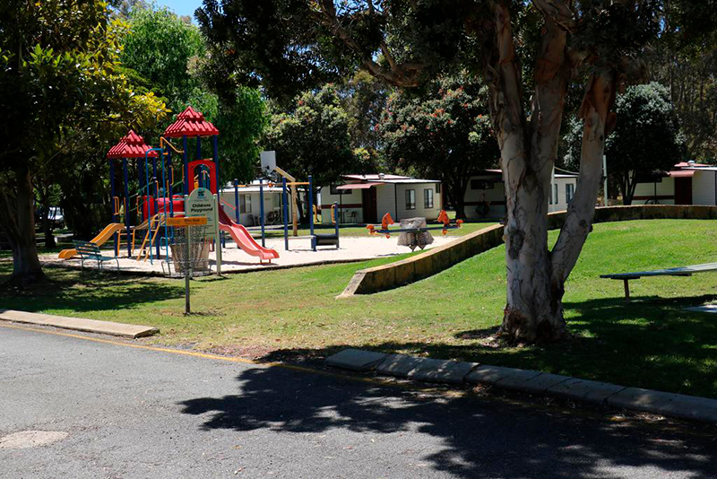 Albany Holiday Park in Western Australia with a disc golf basket beside a children’s playground, cabins, lawns and mature trees.