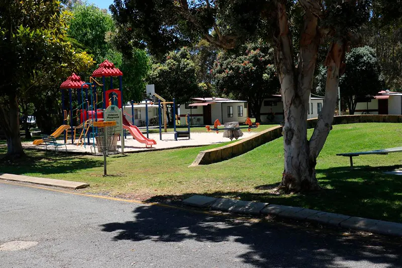 Albany Holiday Park in Western Australia with a disc golf basket beside a children’s playground, cabins, lawns and mature trees.