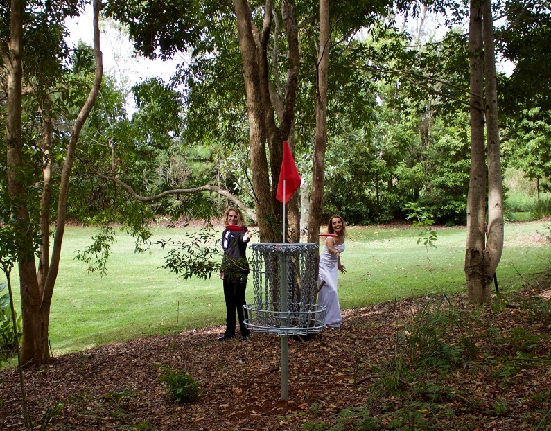 Guests playing disc golf beside a basket at Clouds Eco Resort in Queensland, surrounded by trees and landscaped parkland.